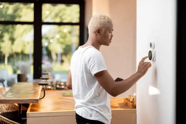 Latin man choosing temperature on thermostat. Young focused guy pushing button on smart home system. Concept of modern domestic lifestyle. Interior of kitchen in modern apartment.