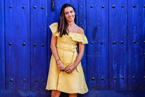 Self-confident and happy woman looking at the camera on a blue background. Charming female in trendy dress leaning on shabby wooden doors of building while looking at camera and relaxing during weekend in city