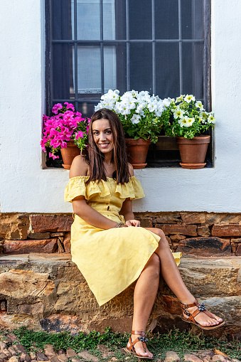 Beautiful woman seated in front of a pretty window with colorful flowers. Woman wearing yellow summer dress smiling at camera.