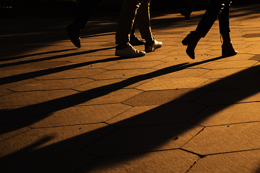 Silhouettes of people legs walking on city street at evening time. Long shadows in golden evening sun. City life.