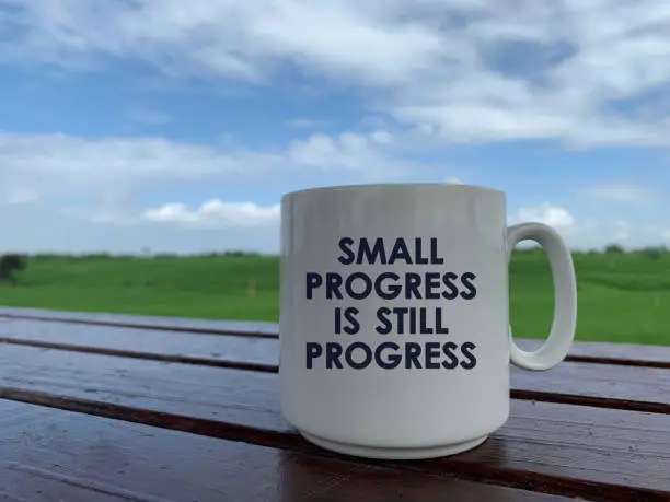 Motivational quote on white a mug - Small progress is still progress. Monday coffee concept with coffee cup on the wooden table with bright summer blue sky clouds and green field background.