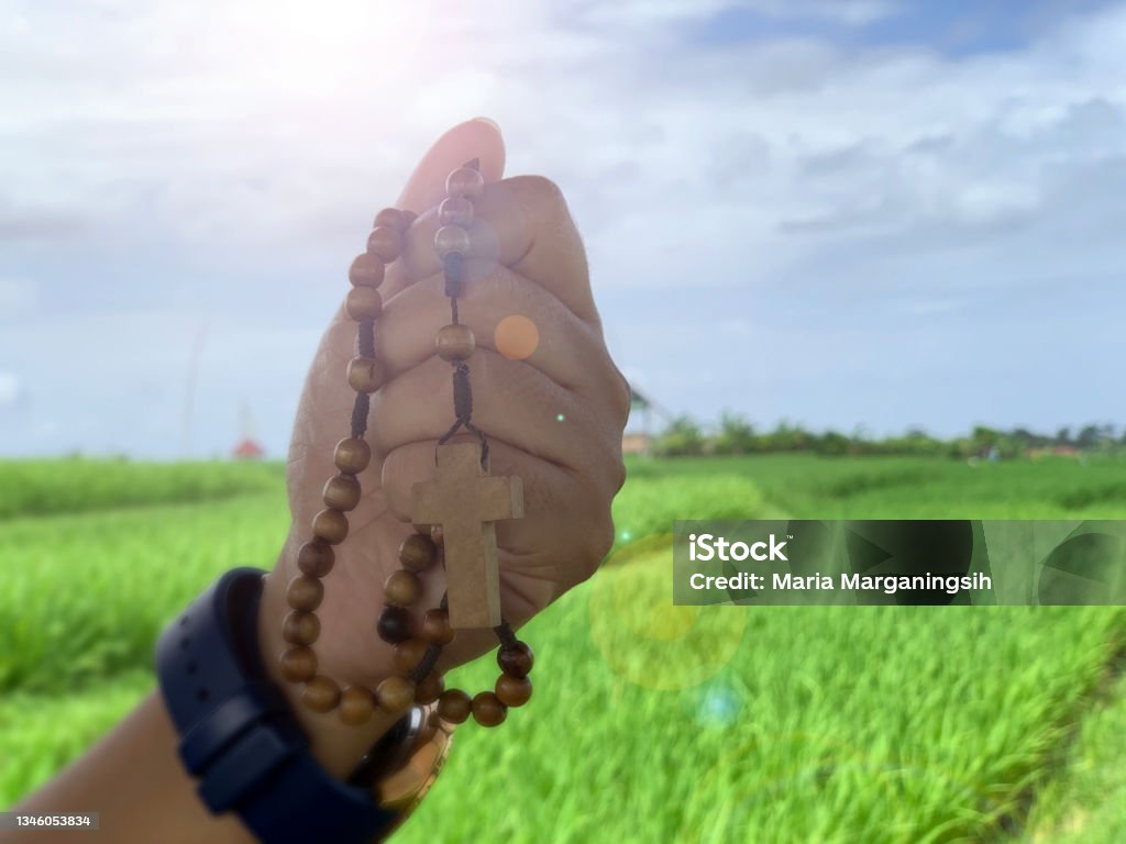 Wooden Rosary Beads in Hand with Jesus Christ holy cross crucifix on bright blue sky light over the green field background. Rosary in hand. Person holding wooden rosary in hands with Jesus Christ holy cross crucifix and light on nature blue sky over green field background. Faith believe in God concept. Catholic symbol. Religion Stock Photo