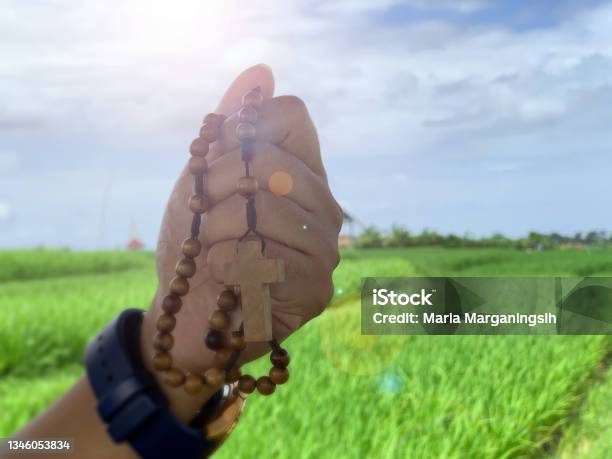 Wooden Rosary Beads In Hand With Jesus Christ Holy Cross Crucifix On Bright Blue Sky Light Over The Green Field Background Stock Photo - Download Image Now