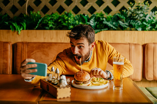 Spontaneous photo of a handsome looking bearded young adult man, sitting at the pub and having a cheeseburger with french fries, and making a selfie wide opening his eyes and mouth next to it. Ready to eat! He’s wearing an ocher sweater over a shirt, looking happy and excited