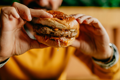 Close up image of an unrecognizable man in yellow holding a fresh burger with both hands while sitting in the restaurant by himself. Focus on a burger. Unhealthy eating concept.