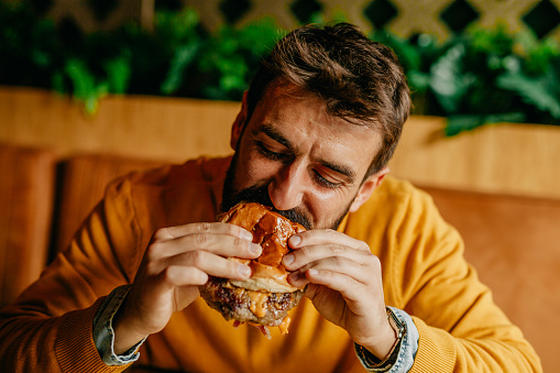 Spontaneous portrait of a handsome bearded man eating a cheeseburger delighted and with enjoyment. He’s elegant casually dressed, wearing an ocher sweater over a jeans shirt, his eyes are closed