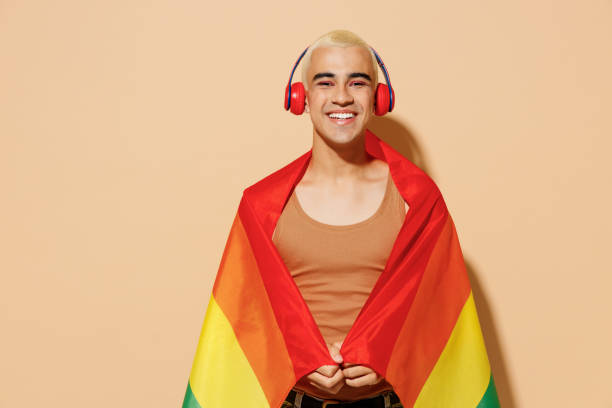 joven divertido sonriente rubio latino gay con maquillaje en camisa de tanque beige auriculares envueltos en bandera arcoíris escuchar música aislada en luz simple estudio de fondo ocre personas concepto de estilo de vida lgbt. - gay pride flag audio fotografías e imágenes de stock