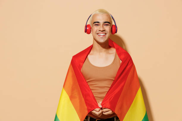 молодой веселый улыбающийся блондин латиноамериканский гей с макияжем в бежевой рубашке наушники, обернутые в радужный флаг, слушает музы� - gay pride flag audio стоковые фото и изображения