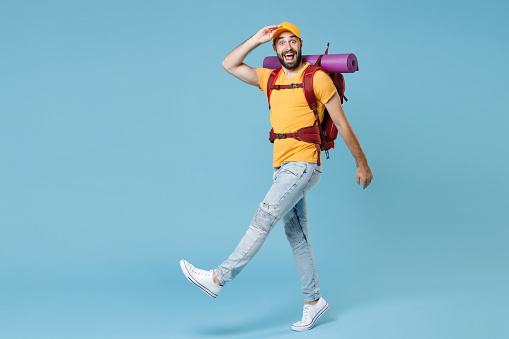 Full length portrait excited traveler man with backpack isolated on blue background. Tourist traveling on weekend. Tourism discovering hiking concept. Hold hand at forehead looking far away distance