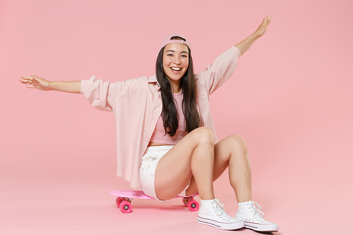 Full length portrait of cheerful young asian woman girl in casual clothes, cap isolated on pastel pink background studio portrait. People lifestyle concept. Sit on skateboard, rising spreading hands