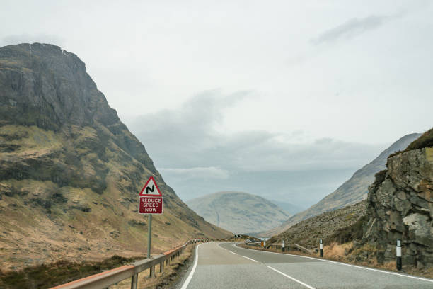 Main Road through Glencoe Mountains with Reduce Speed Road Safety Sign Glencoe is situated between the banks of Loch Leven and the mouth of the famed valley, making it an ideal location for exploring Lochaber, known as the UK's Outdoor Capital. Glen Coe's road leads you right through the centre of an ancient volcano. Follow the Glen Coe Geotrail to learn more about the glen's craggy mountain peaks and how glaciers and violent explosions chiselled them out. Glen Coe is one of Scotland's most gorgeous and otherworldly locations. It's even been seen in movies like James Bond's Skyfall and various Harry Potter films. glencoe scotland photos stock pictures, royalty-free photos & images