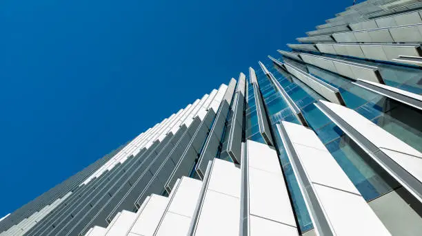 Upward view from street level of a tall modern office building exterior. Against a blue sky. Adjustable external slats to control incoming natural sunlight for internal light and temperature control.