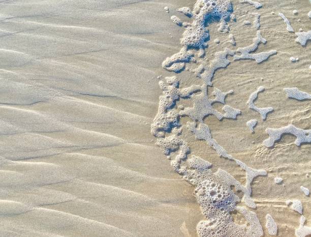 Horizontal high angle view close up of salt water sea foam ocean waves breaking on beach sand creating natural abstract pattern design