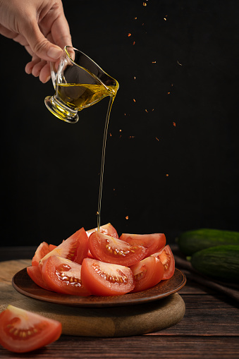 Sliced tomato vegetable served on plate with hand pouring olive oil from glass transparent gravy boat and falling hot chili spices on dark brown wooden table with cucumber against black wall for lunch