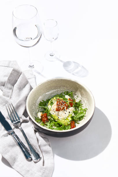 istock Avocado and stracciatella cheese salad. Fancy dinning with Italian salad bowl on white table with simple contemporary decor. Sunlight and harsh shadow still life. Avocado, cheese and tomato salad. 1346110369