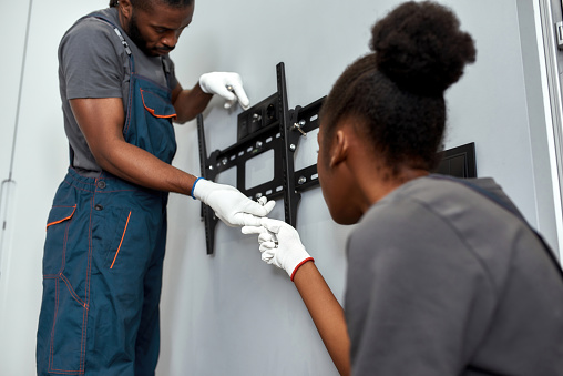 Close view African man and woman during TV wall brackets installation work. Female assistant giving tools to handyman, both in white protective gloves. Team work concept.