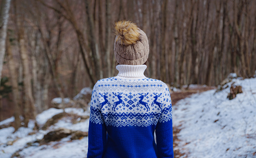Beautiful woman standing among trees in winter forest. Wearing hat and blue sweater. Caucasian Asian female model outside in first snow