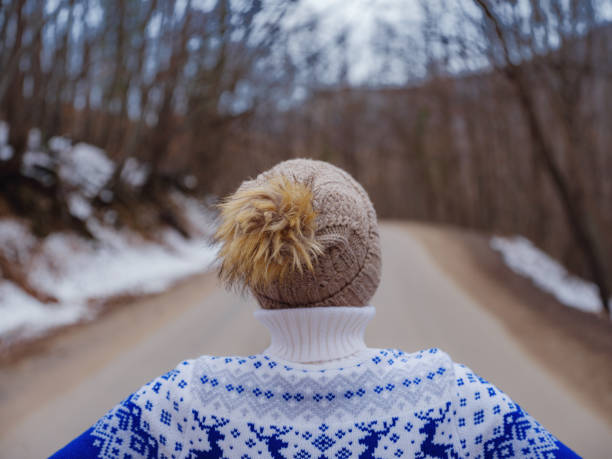 Beautiful woman standing among trees in winter forest. Wearing hat and blue sweater. Caucasian Asian female model outside in first snow