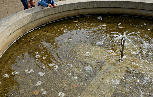 stone sandstone round fountain in the park. built of sandstone filled with water. lined with a light threshing gravel path with the edge of granite cubes, two boys pour over the edge to the water