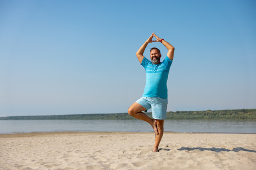 Cheerful mid adult man in blue shorts and T-shirt exercising on sandy beach, arms raised, hands in steeple, standing on one leg, the other bent inwards, yoga tree pose Vrksasana