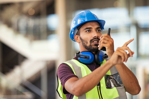 Construction supervisor using walkie talkie Young mixed race supervisor instructing workers using walkie-talkie at construction site. Contractor wearing yellow vest and blue helmet using walkie talkie to explain employees what to do. Young indian engineer working at construction site. walkie talkie photos stock pictures, royalty-free photos & images