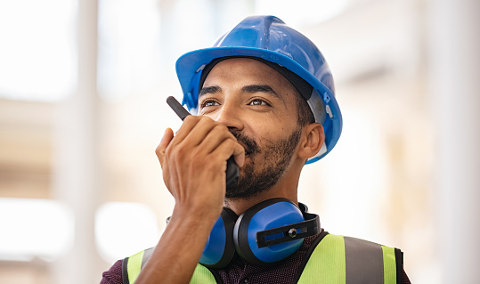 Young middle eastern bricklayer using walkie-talkie at construction site. Smiling indian site manager wearing helmet and yellow vest with safety ear muff while talking through walkie-talkie. Confident supervisor using walkie-talkie, giving instructions at construction site.