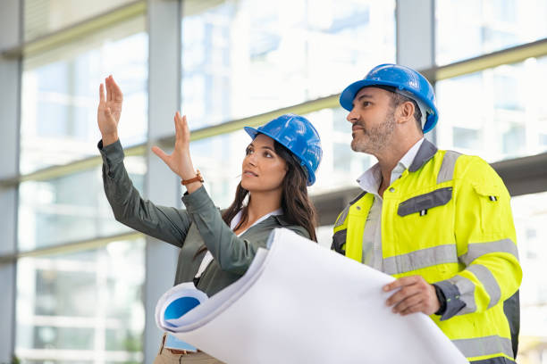 Architect and contractor imagine the building under construction Young woman architect explaining blueprint to supervisor wearing safety vest at construction site. Mid adult contractor holding blueprint and understanding manager vision at construction site. Smiling engineer with hardhat on head talking to contractor while standing in building in construction process. foreperson stock pictures, royalty-free photos & images