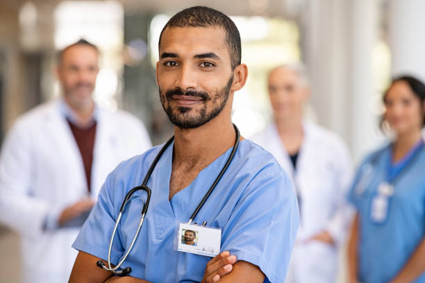 Young confident male nurse looking at camera Portrait of smiling middle eastern man nurse in uniform and stethoscope looking at camera. Young doctor smiling while standing in hospital corridor with health care team in background. Successful indian surgeon smiling in front of his multiethnic medical team. medical scrubs stock pictures, royalty-free photos & images