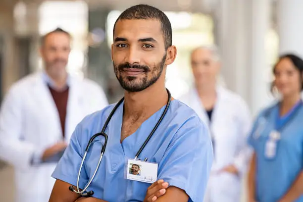 Portrait of smiling middle eastern man nurse in uniform and stethoscope looking at camera. Young doctor smiling while standing in hospital corridor with health care team in background. Successful indian surgeon smiling in front of his multiethnic medical team.