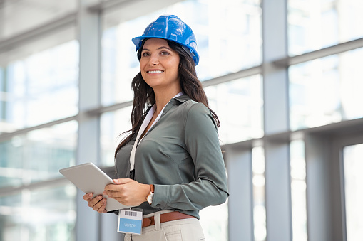 Beautiful mid adult woman architect wearing blue hardhat at construction site while working on digital tablet. Supervisor wearing safety helmet while working in a building site. Successful and proud inspector looking away with copy space.