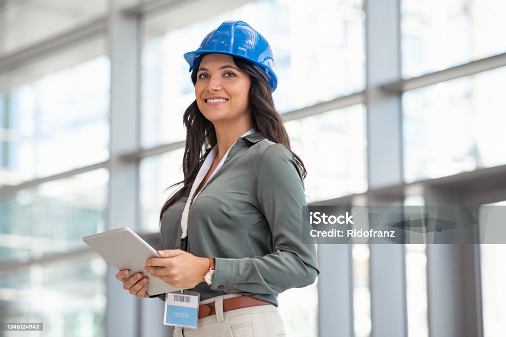 Architect working on digital tablet at construction site Beautiful mid adult woman architect wearing blue hardhat at construction site while working on digital tablet. Supervisor wearing safety helmet while working in a building site. Successful and proud inspector looking away with copy space. Engineer Stock Photo