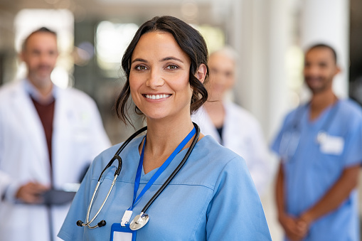 Portrait of smiling young nurse in uniform smiling with healthcare team in background. Successful team of doctor and nurses smiling. Beautiful and satisfied healthcare worker standing in private clinic and looking at camera with pride.