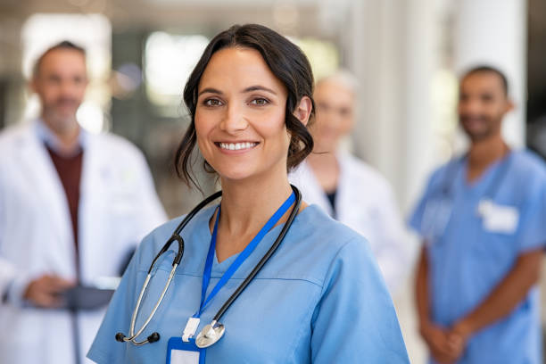 Portrait of friendly nurse smiling at hospital Portrait of smiling young nurse in uniform smiling with healthcare team in background. Successful team of doctor and nurses smiling. Beautiful and satisfied healthcare worker standing in private clinic and looking at camera with pride. nurse stock pictures, royalty-free photos & images