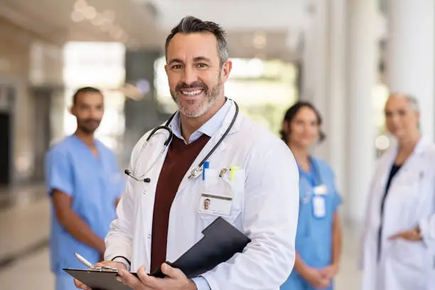 Portrait of handsome mature doctor wearing labcoat and stethoscope reading patient case file in hospital corridor. Successful head physician standing in hospital hallway and looking at camera with his medical team in background. Reliable general practitioner in career with his healthcare staff.