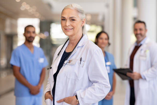 Senior confident doctor with his medical staff at hospital Portrait of mature female doctor on hospital corridor with hands in labcoat pockets and stethoscope. Confident general practitioner standing in hospital hallway with her healthcare team in background. Successful head physician in private clinic smiling and looking at camera. female doctor stock pictures, royalty-free photos & images