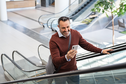 Smiling mid adult business man smiling while reading message on smartphone at airport. Successful senior casual man going up the escalator using smart phone while going to work. Businessman doing video call while standing in commercial building and using wireless earbuds.