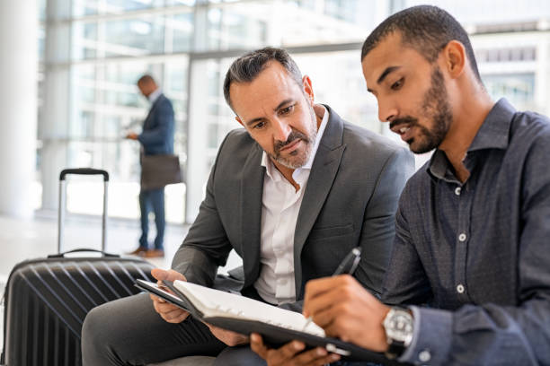 Multiethnic businesspeople at airport talking Two multiethnic businesspeople sitting at departure lounge at the airport discussing work while making notes in agenda. Indian business man working at airport with worried manager while waiting in lounge their connection. Successful businessman discussing during a business travel. leaving a tip stock pictures, royalty-free photos & images