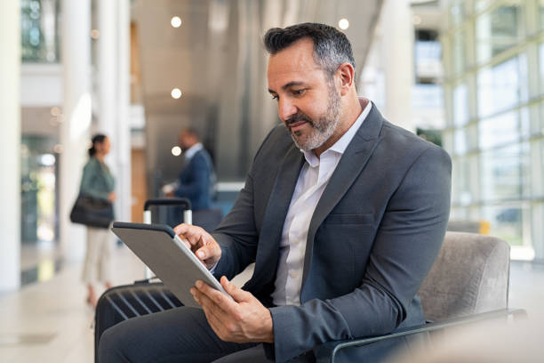 Mature businessman using digital tablet at departure lounge at the airport Successful business man sitting in airport lounge working on digital tablet. Mature businessman using digital tablet while sitting and waiting for boarding at international airport gate. Mid adult businessman in formalwear sitting in lounge of modern airport and working. mature businessman stock pictures, royalty-free photos & images