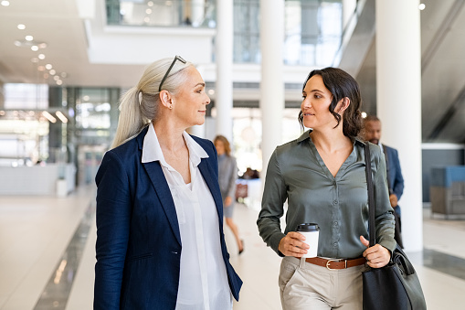 Two business women walking and talking in modern office. Two successful women manager discussing work while entering office building. Successful mature business woman talking with her assistant while going to work.