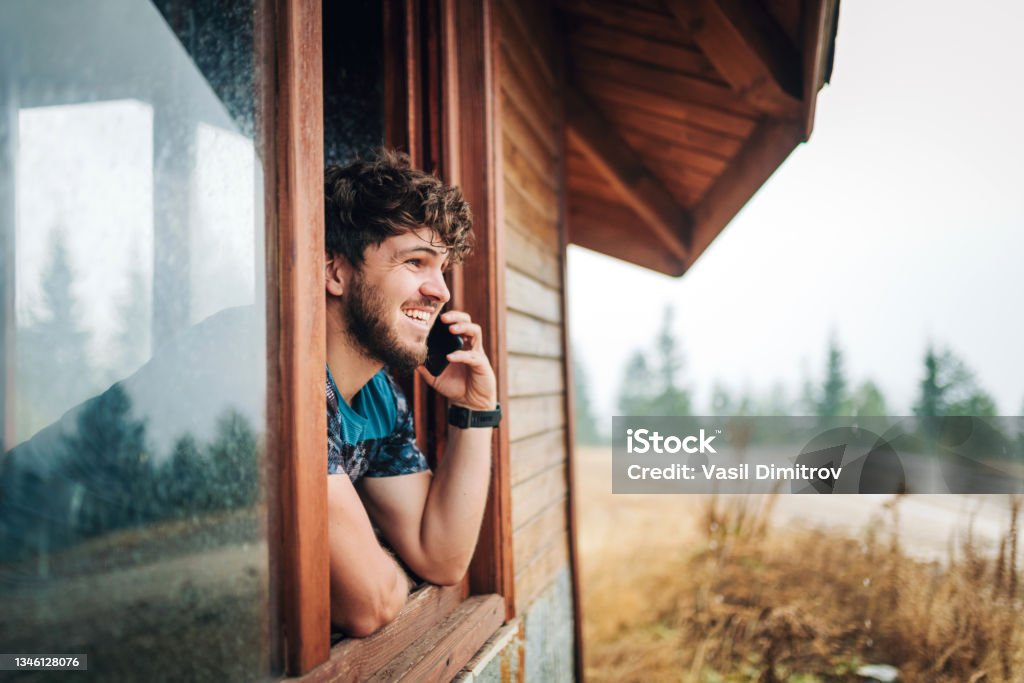 Tell your friends about your adventures. Young man enjoying a cold autumn day in the mountain and talking on the phone. Log Cabin Stock Photo