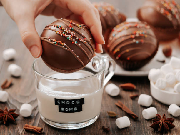 Chocolate cocoa bomb or ball in hand near milk cup Chocolate cocoa bomb in hand near glass cup with plant-based milk and choco bomb text. Ball made from milk chocolate with marshmallow. Stylish orange toned image of trendy winter hot chocolate drink bomb stock pictures, royalty-free photos & images