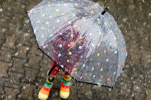 Little toddler girl playing with big umbrella on rainy day. Happy positive child running through rain, puddles. Preschool kid with rain clothes and rubber boots. Children activity on bad weather day