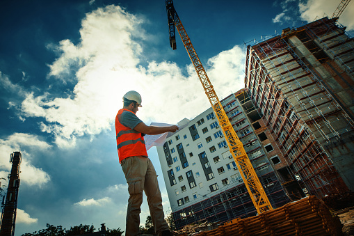 Closeup low angle view construction engineer standing in front of a building. He's doing some calculation and inspection by looking at construction plan