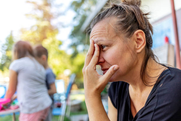 Stressed depressed young mother sits outside in a playground with her kids playing behind her. Stressed depressed young mother sits outside in a playground with her kids playing behind her. Single frustrated woman hold her head with hands sitting on chair with playful kids on a background. single mother stock pictures, royalty-free photos & images