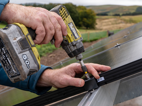 Freestanding solar pv panels being fitted for domestic use, by an engineer using a handheld power tool. The individual panels are being screwed onto metal posts and a metal frame, set on a concrete base. They are positioned to catch maximum sunlight to create electricity from a sustainable renewable energy source.