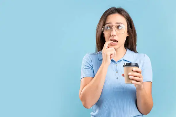 Confused thoughtful young woman puts finger in her mouth, holds paper cup with coffee, wearing casual t-shirt and eyeglasses