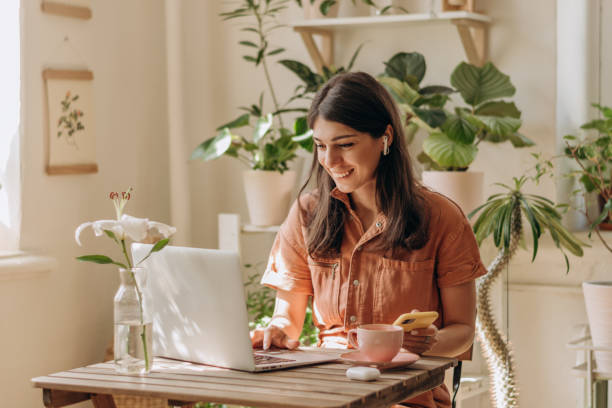 positive junge mixed-race-frau mit laptop und smartphone zu hause. gemütliches wohninterieur mit zimmerpflanzen. remote-arbeit, business, freelance, online-shopping, e-learning, großstadt-dschungel-konzept - 1 stock-fotos und bilder