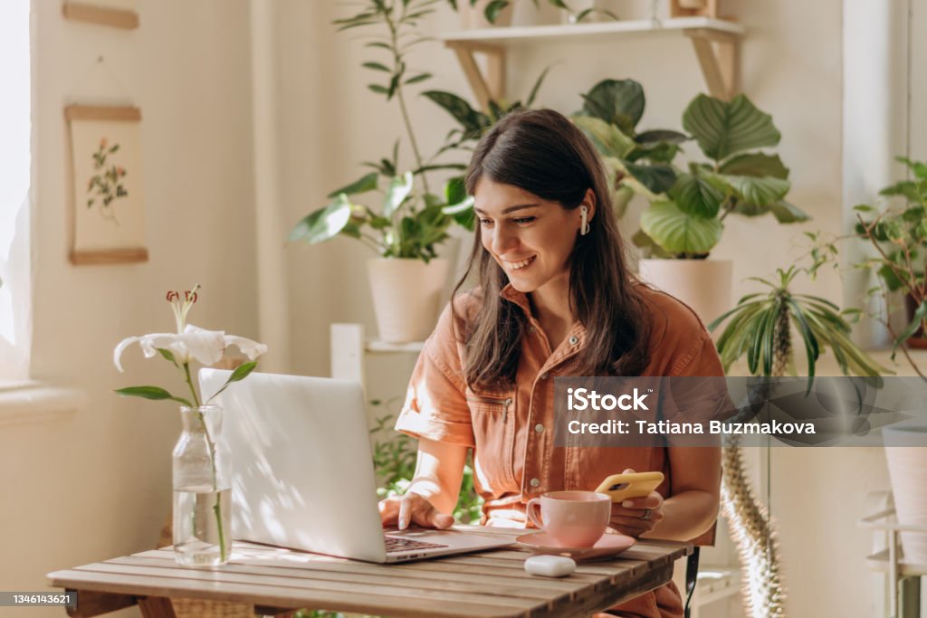 Positive young mixed race woman using a laptop and smartphone at home.Cozy home interior with indoor plants.Remote work, business,freelance,online shopping,e-learning,urban jungle concept Positive young mixed race woman using a laptop and smartphone at home.Cozy home interior with indoor plants.Remote work, business,freelance,online shopping,e-learning,urban jungle concept. Working At Home Stock Photo