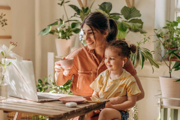 Positive young woman of mixed race with her little daughter is sitting at home at her desk and using a laptop.Remote work at home, freelance, working mom,online education. stock photo