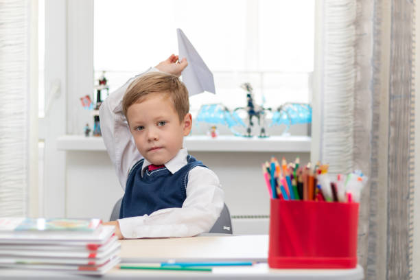 A cute first-grader boy in a school uniform at home while isolated at his desk makes a paper airplane during recess. Selective focus
